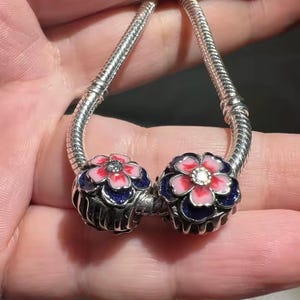 May include: Two silver flower charms with pink and blue enamel details. Each charm features a pink flower with a central gem, set against a dark blue background. The charms are attached to a silver bracelet.