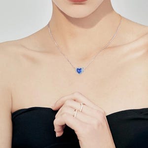May include: A silver necklace with a heart-shaped pendant. The pendant features a vibrant blue center stone surrounded by a halo of smaller, clear stones. The necklace is worn around the neck.