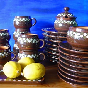 Può includere: Un set di tazze in ceramica marrone con pois bianchi, una zuccheriera in ceramica marrone con coperchio e una pila di piatti in ceramica marrone con pois bianchi. Le tazze e i piatti sono disposti su una superficie di legno con limoni in primo piano.