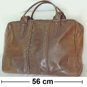 May include: A brown leather travel bag with a top handle and a shoulder strap. The bag has a zipper closure and is decorated with metal studs. The bag measures 56 cm wide and 58 cm tall.