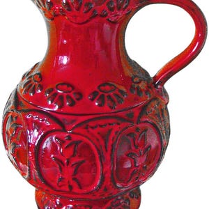 Può includere: Una brocca in ceramica rossa con manico e beccuccio. La brocca ha un corpo bombato con motivi floreali e geometrici in rilievo. Il manico è arcuato e il beccuccio leggermente svasato. La brocca è di un rosso intenso e lucido con accenti scuri.