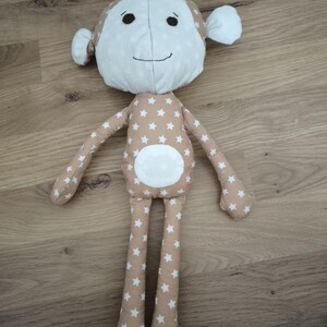 Op de afbeelding: Een bruin pluche apen speelgoed met witte sterren op zijn lichaam en een witte cirkel op zijn buik. De aap heeft een wit gezicht met zwarte ogen en een gestikte glimlach.