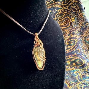 May include: A handcrafted pendant necklace featuring a polished, iridescent gemstone wrapped in copper wire. The gemstone displays shades of green, yellow, and blue. The pendant hangs from a thin, brown cord, creating a unique and artistic piece of jewelry.