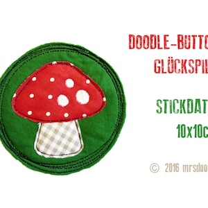 May include: A green felt circle with a red and white mushroom applique. The mushroom has white polka dots and a checkered stem. The text "DOODLE-BUTTON GLÜCKSPILZ STICKDATEI 10x10cm © 2016 mrsdoodle" is printed below the circle.