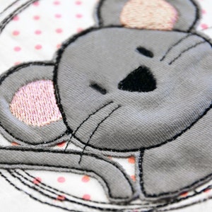 Pode incluir: Um rato cinzento bordado com orelhas cor-de-rosa e um nariz preto, sobre um fundo branco com bolinhas cor-de-rosa.