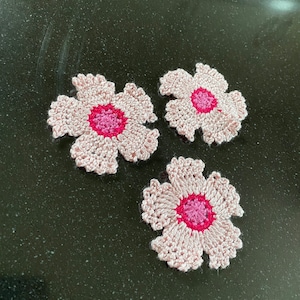 Könnte beinhalten: Drei gehäkelte Blumenapplikationen. Jede Blume hat ein hellrosa Blütenblattmuster mit einem dunkleren rosa Zentrum. Die Blumen sind auf einer dunklen, gesprenkelten Oberfläche angeordnet.