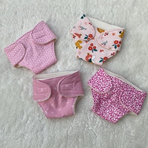 Könnte beinhalten: Vier Stoffwindeln in verschiedenen rosa Mustern werden auf einer weißen, flauschigen Oberfläche präsentiert. Eine Windel hat weiße Tupfen, eine andere ein Blumendesign mit einem Eichhörnchen, und die anderen haben Blatt- und Blumenmuster.
