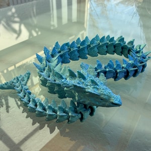 Könnte beinhalten: Blaues, bewegliches Drachenfigur mit strukturierter Oberfläche. Der Drache hat einen langen, segmentierten Körper mit stacheligen Vorsprüngen entlang seines Rückens und Schwanzes. Der Drache ist auf einer reflektierenden Oberfläche abgebildet.