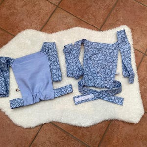 Könnte beinhalten: Ein Babytrageset mit hellblauem Blumenmuster auf einer weißen, flauschigen Oberfläche. Das Set enthält eine Trage, Träger und einen Hüftgurt. Die Trage hat eine einfarbige hellblaue Platte.