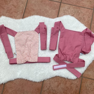 Könnte beinhalten: Zwei Puppenträger in rosa und weiß mit Blumen- und Tupfenmustern. Die Träger sind aus Stoff und haben Riemen und Verschlüsse. Einer hat eine florale Mitte, der andere ein Tupfenmuster.