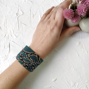 以下が含まれることがあります： 青、緑、黒のパターンでビーズで作られたブレスレット。ブレスレットは手首につけられています。