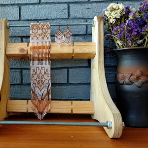 May include: A wooden jewelry stand with a brown beaded necklace and a pair of white beaded earrings. The stand has a wooden base and two shelves. The necklace and earrings are displayed on the top shelf. The stand is in front of a brick wall.