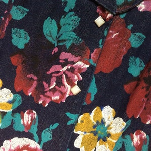 May include: A dark blue denim shirt with a floral print in shades of pink, red, white, and yellow. The shirt has a button-up closure and features silver-toned buttons.