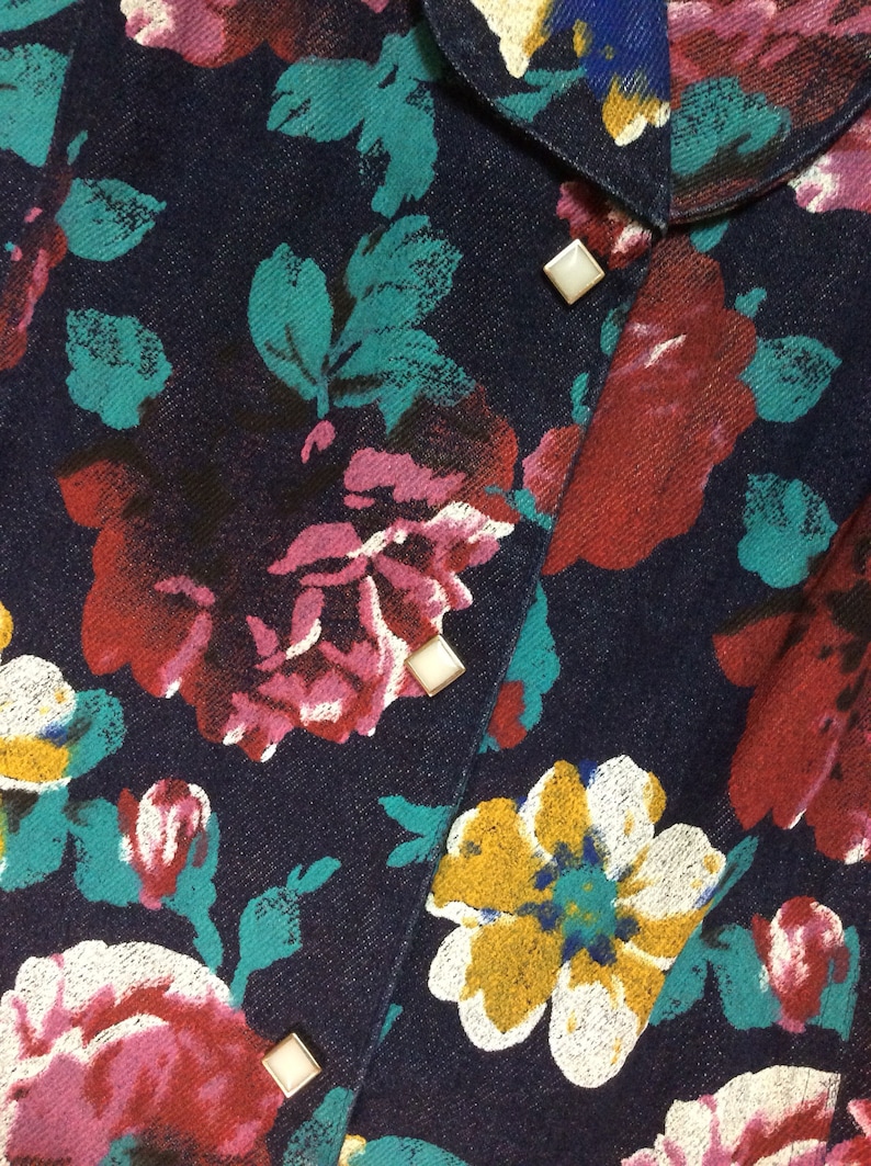 May include: A dark blue denim shirt with a floral print in shades of pink, red, white, and yellow. The shirt has a button-up closure and features silver-toned buttons.