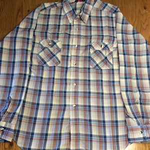 Puede incluir: Una camisa de cuadros de manga larga con cierre de botones. La camisa es blanca con cuadros azules, rojos y naranjas. La camisa tiene dos bolsillos en el pecho con cierre de botones.