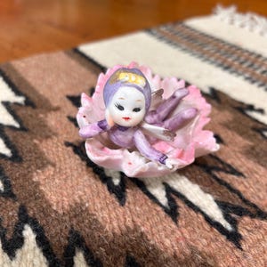 May include: A porcelain figurine of a small fairy with a purple hood and outfit, resting inside a large, open pink flower petal. The fairy has painted facial features, including dark eyelashes and red lips, and small wings extending from the back.