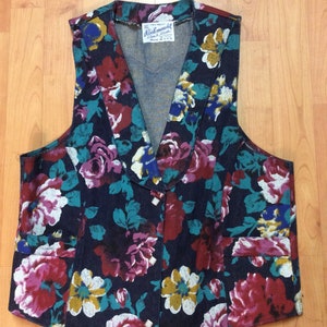 May include: A dark blue denim vest with a floral print featuring red, white, yellow, and teal flowers. The vest has a V-neck and a button closure. The label reads "Rockmount Ranch Wear Made in U.S.A."