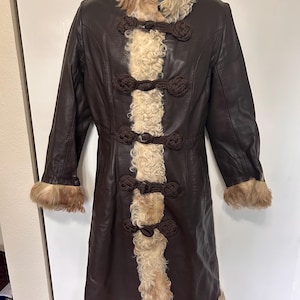 May include: A long brown leather coat with a white fur collar and trim. The coat has a unique design with a vertical strip of white fur down the front and brown rope-like closures.