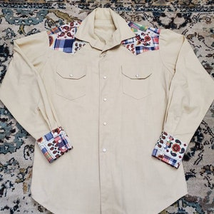 May include: A beige long-sleeved button-down shirt with a patchwork design on the shoulders and cuffs. The patchwork includes paisley, plaid, and floral patterns in shades of red, blue, green, and white.