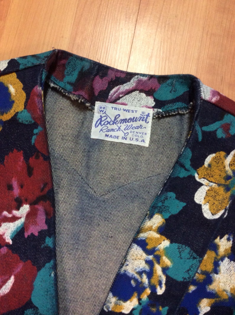 May include: A dark blue denim jacket with a floral print. The jacket has a label that reads "Rockmount Ranch Wear Denver Colo. Made in U.S.A."