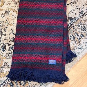 Op de afbeelding: Rode en groene gepatroneerde deken met een blauwe franje en een blauw label met de tekst "Remarkable Products".