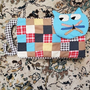 May include: A patchwork cat pillow with a blue head and a brown and white houndstooth tail. The cat's head is a separate pillow with a stitched-on face.