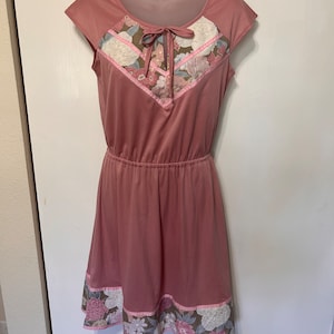 May include: A pink sleeveless dress with a floral print on the neckline and hem. The dress has a tie at the neckline and a fitted waist.