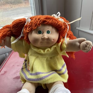 May include: A vintage Cabbage Patch Kid doll with orange yarn hair styled in pigtails, white ribbons, and green eyes. The doll wears a yellow knit dress with purple stripes and white socks. The doll is sitting on a red surface.