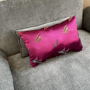 May include: A magenta rectangular decorative pillow with embroidered butterflies in shades of brown and gold. The pillow is placed on a gray sofa, offering a pop of color and a touch of nature-inspired design to the living space.