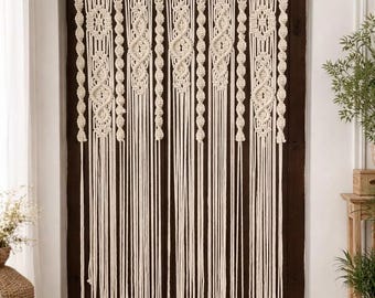 May include: Cream-colored macrame door curtain. The curtain features intricate woven patterns at the top and along the vertical strands. The design includes geometric shapes and knotted details, creating a bohemian aesthetic. The curtain hangs in front of a dark brown door.