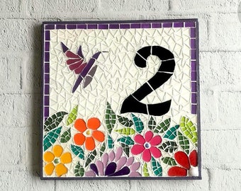 Puede incluir: Placa de número de casa de mosaico con el número 2 en negro, rodeada de flores coloridas y un colibrí morado. La placa tiene un borde morado.