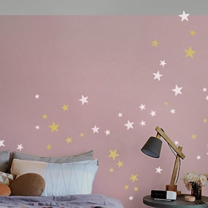 Könnte beinhalten: Eine rosa Wand mit weißen und goldenen Sternen, ein Bett mit Kissen und einer Decke sowie ein Nachttisch mit einer Lampe und einer Uhr.
