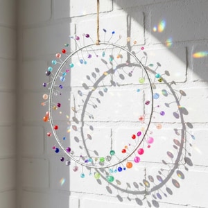 Könnte beinhalten: Ein dekorativer, kreisförmiger Wandschmuck mit einem silbernen Metallrahmen. Der Rahmen ist mit bunten, irisierenden Perlen in Rot-, Orange-, Gelb-, Grün-, Blau- und Lilatönen verziert. Das Stück hängt an einer Naturfaserschnur vor einer weißen Backsteinmauer.