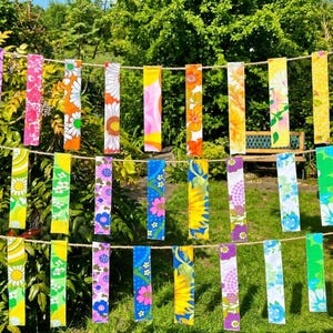 以下が含まれることがあります： カラフルな長方形の布のストリップが紐に吊るされており、花柄が施されています。 ストリップには、ピンク、オレンジ、黄色、青の色合いで、デイジーやヒマワリなどのデザインが施されています。 背景は緑豊かな庭で、明るい雰囲気を作り出しています。