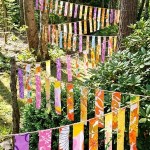 以下が含まれることがあります： 木々の間に張られたロープから、花柄や抽象的な模様のカラフルな布のストリップが吊り下げられています。 ストリップは紫、ピンク、オレンジ、黄色の色合いで、活気のある屋外装飾を作り出しています。 緑豊かな森の中のシーンです。