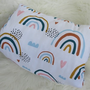 Könnte beinhalten: Rechteckiges Kissen mit weißem Hintergrund und einem sich wiederholenden Muster aus Regenbogen in verschiedenen Farben, darunter Türkis, Braun und Gold. Das Design zeigt auch kleine rosa Herzen, blaue Wolken und schwarze Punkte.