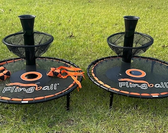 Può includere: Due giochi di trampolino Flingball neri e arancioni con reti nere e palle arancioni. Ogni gioco ha una base nera con un palo nero e una rete nera. I giochi sono su una superficie erbosa verde.