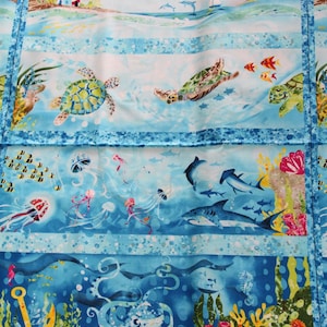 Könnte beinhalten: Ein blauer und weißer Stoff mit einem sich wiederholenden Muster aus Meereslebewesen. Das Muster umfasst einen Leuchtturm, Delfine, Meeresschildkröten, Quallen, Haie und einen Oktopus. Der Stoff ist perfekt zum Quilten, Nähen und Basteln.