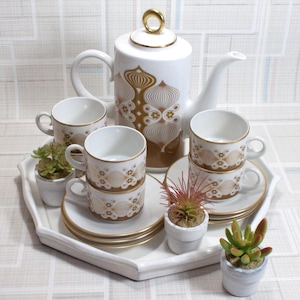 Könnte beinhalten: Eine weiße Teekanne und vier Teetassen mit einem braunen und goldenen Blumendesign. Die Teekanne hat einen goldenen Griff und einen goldenen Knopf auf dem Deckel. Die Teetassen sind auf einem weißen Unterteller mit goldenen Rand angeordnet.