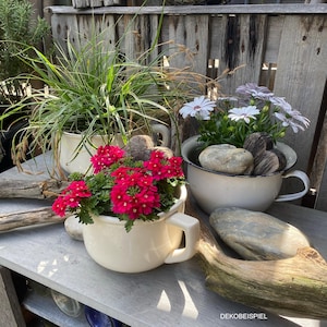 Op de afbeelding: Drie crèmekleurige emaille plantenbakken met handvatten, gevuld met levendige planten en bloemen. Eén bevat felrode bloemen, een andere witte en paarse madeliefjes en de derde lang groen gras. Decoratieve stenen en drijfhout dragen bij aan de rustieke uitstraling.