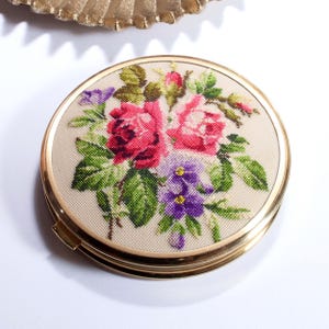 Könnte beinhalten: Ein runder, goldfarbener Kompaktspiegel mit einem floralen Kreuzstichdesign. Das Design zeigt rosa Rosen, lila Veilchen und grüne Blätter auf beigem Hintergrund. Der Kompaktspiegel hat eine Klappöffnung und ein glänzendes Finish.