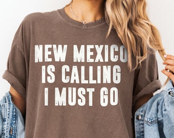 May include: A brown t-shirt with the text "NEW MEXICO IS CALLING I MUST GO" in white distressed lettering.