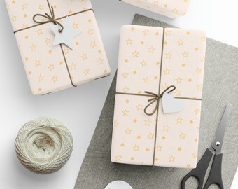 Puede incluir: Tres regalos envueltos con papel estampado de estrellas, atados con hilo marrón y etiquetas blancas en forma de estrella. También hay un ovillo de hilo, tijeras y etiquetas en blanco, lo que sugiere un tema de manualidades o envoltura de regalos.