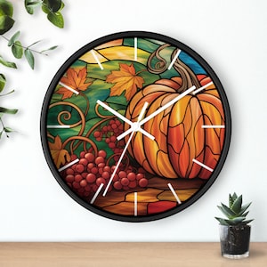 Könnte beinhalten: Eine runde Wanduhr mit schwarzem Rahmen. Das Zifferblatt zeigt eine Buntglas-Illustration eines Kürbisses, Trauben und Herbstblättern in Orange, Rot und Grün. Weiße Zeiger zeigen die Uhrzeit an.