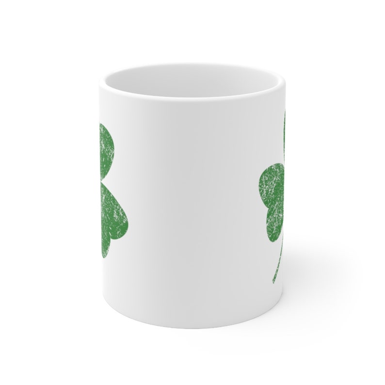May include: White ceramic mug featuring a distressed green shamrock design. The shamrock is a symbol of luck and is commonly associated with St. Patrick's Day. The mug is a standard size and shape, suitable for hot or cold beverages.