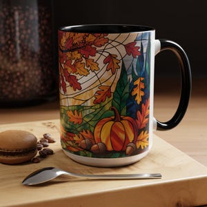 May include: A white ceramic mug with a black handle and rim. The mug features a colorful autumn design with a pumpkin, fall leaves, and acorns. The mug is on a wooden surface with a spoon and a macaron.