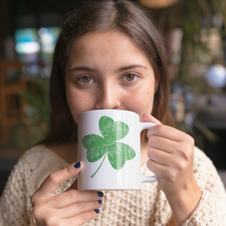 A young woman models an 11-ounce white ceramic coffee mug featuring Sweet Holdings distressed, green shamrock, design, printed on both the front and back sides of the mug.