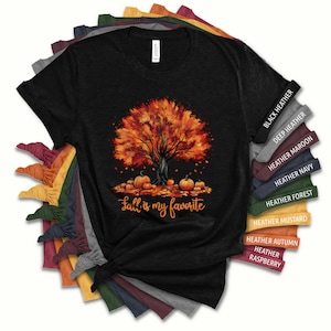 Könnte beinhalten: Ein schwarzes T-Shirt mit einem Aufdruck eines Herbstbaums mit orangefarbenen, roten und gelben Blättern. Kürbisse sind um den Fuß des Baumes verstreut. Der Text "Fall is my favorite" ist unter dem Baum aufgedruckt. Das Shirt ist in verschiedenen Farben erhältlich, darunter Schwarz meliert, Dunkel meliert, Rotbraun meliert, Marineblau meliert, Waldgrün meliert, Senf meliert, Herbst meliert und Himbeer meliert.