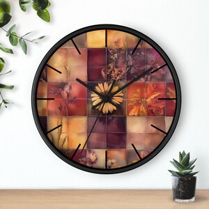 May include: A round wall clock with a black frame. The clock face features a mosaic of squares in shades of orange, yellow, and burgundy, with floral and botanical imagery. A large yellow flower is at the center. Black clock hands indicate the time.