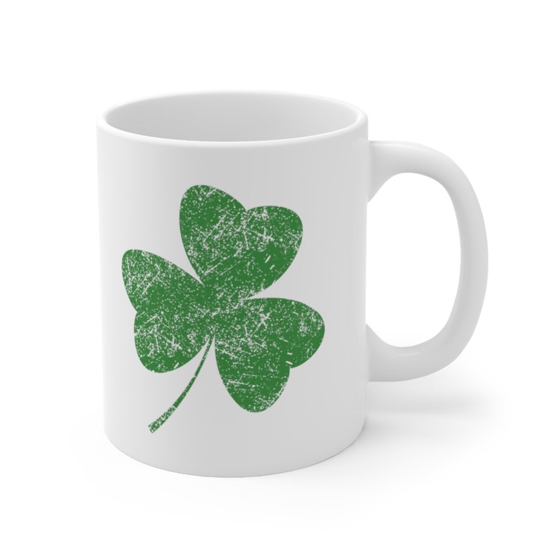 An 11-ounce white ceramic coffee mug featuring Sweet Holdings distressed, green shamrock, design, printed on both the front and back sides of the mug.